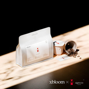 A promotional image showing a white coffee bag and XBloom xpods coffee capsules on a wooden surface with a dark shadow background, showcasing the 'xbloom x algebraist' collaboration text.