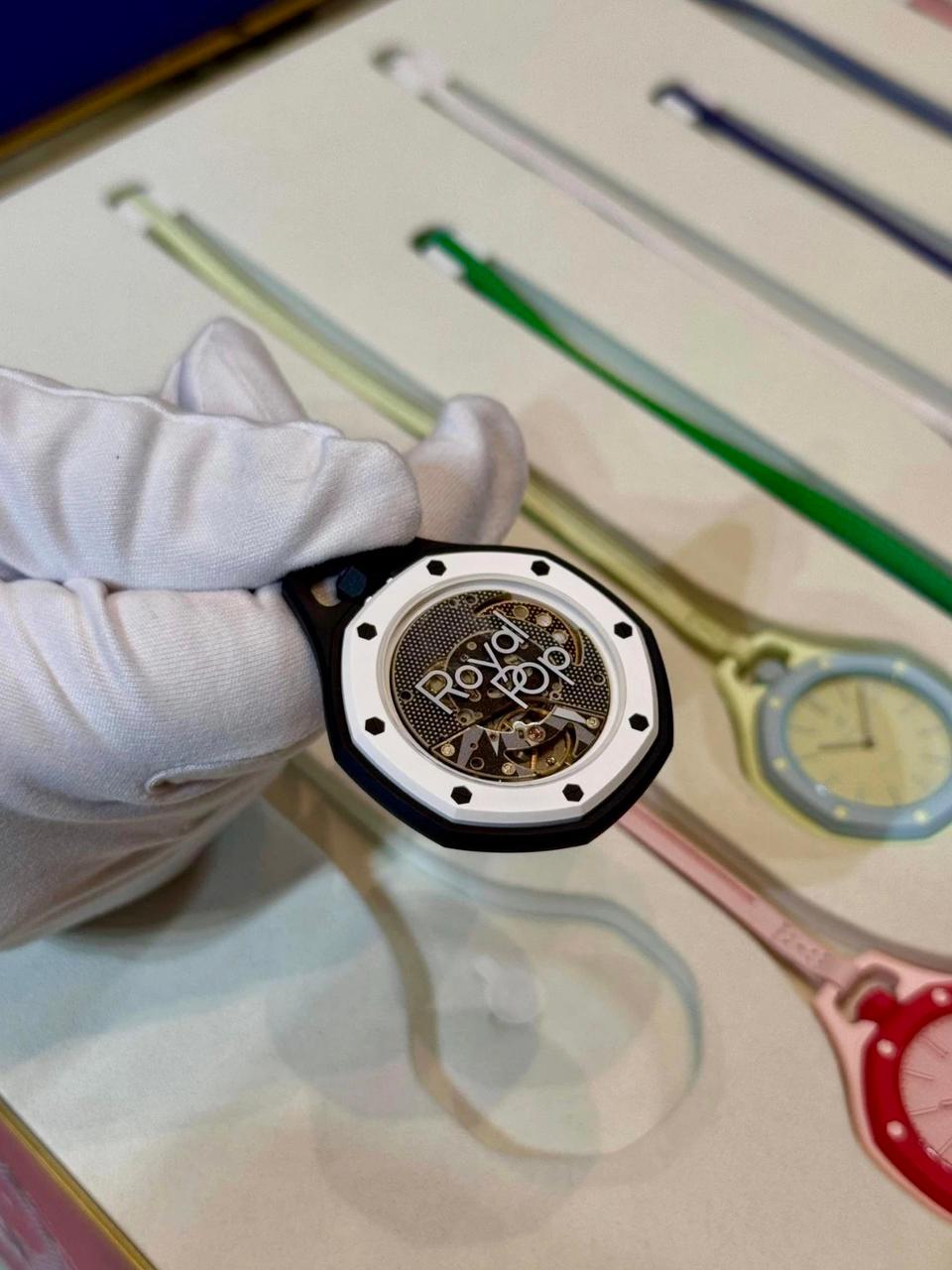 A close-up of a gloved hand holding a black bioceramic Royal Pop pocket watch with a skeleton dial, displaying the internal mechanical movement against a background of colorful watches.